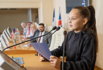 Cute schoolgirl with clipboard speaking in microphone while making report by tribune against row of foreign delegates